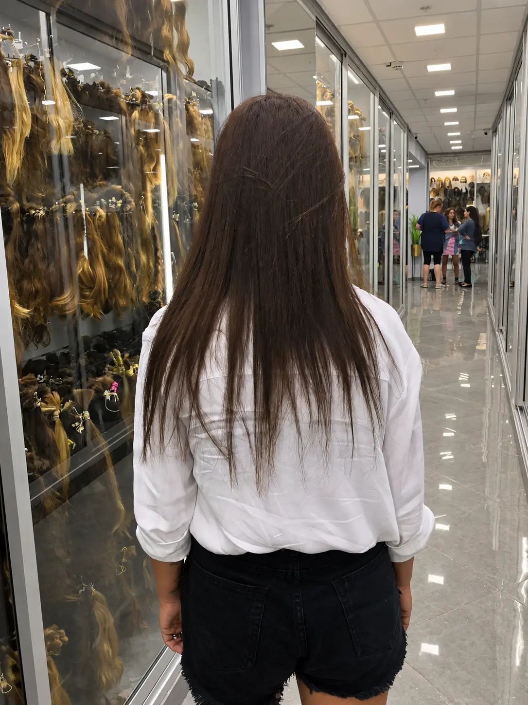 הארכת שיער לפני: לקוחה אמיתית מהגב עם שיער חום חלק עד אמצע הגב, דק ודליל בקצוות, במסדרון הסטודיו מול ויטרינות שיער
